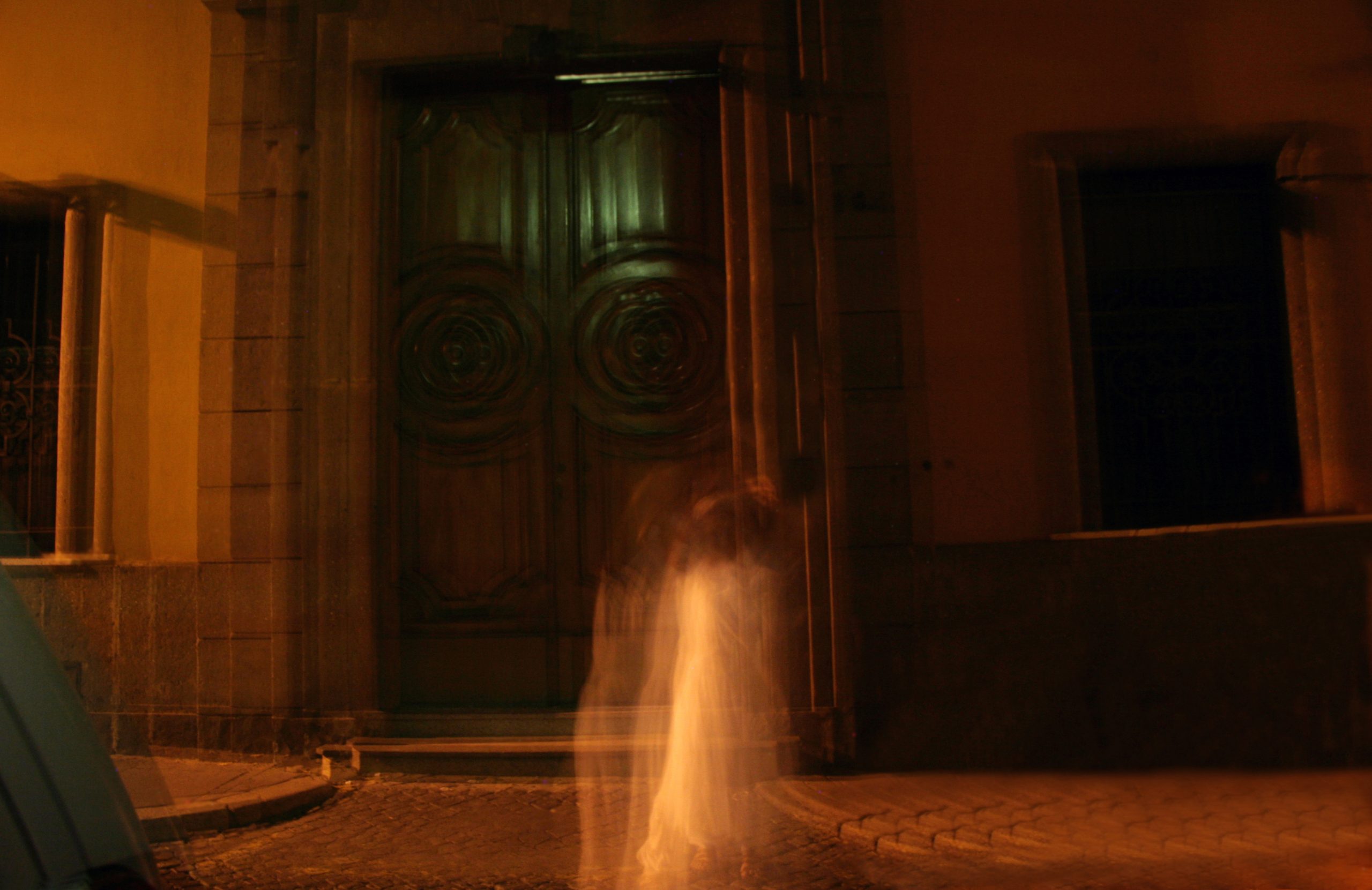 Blurred figure in white dress in front of large wooden doors at night